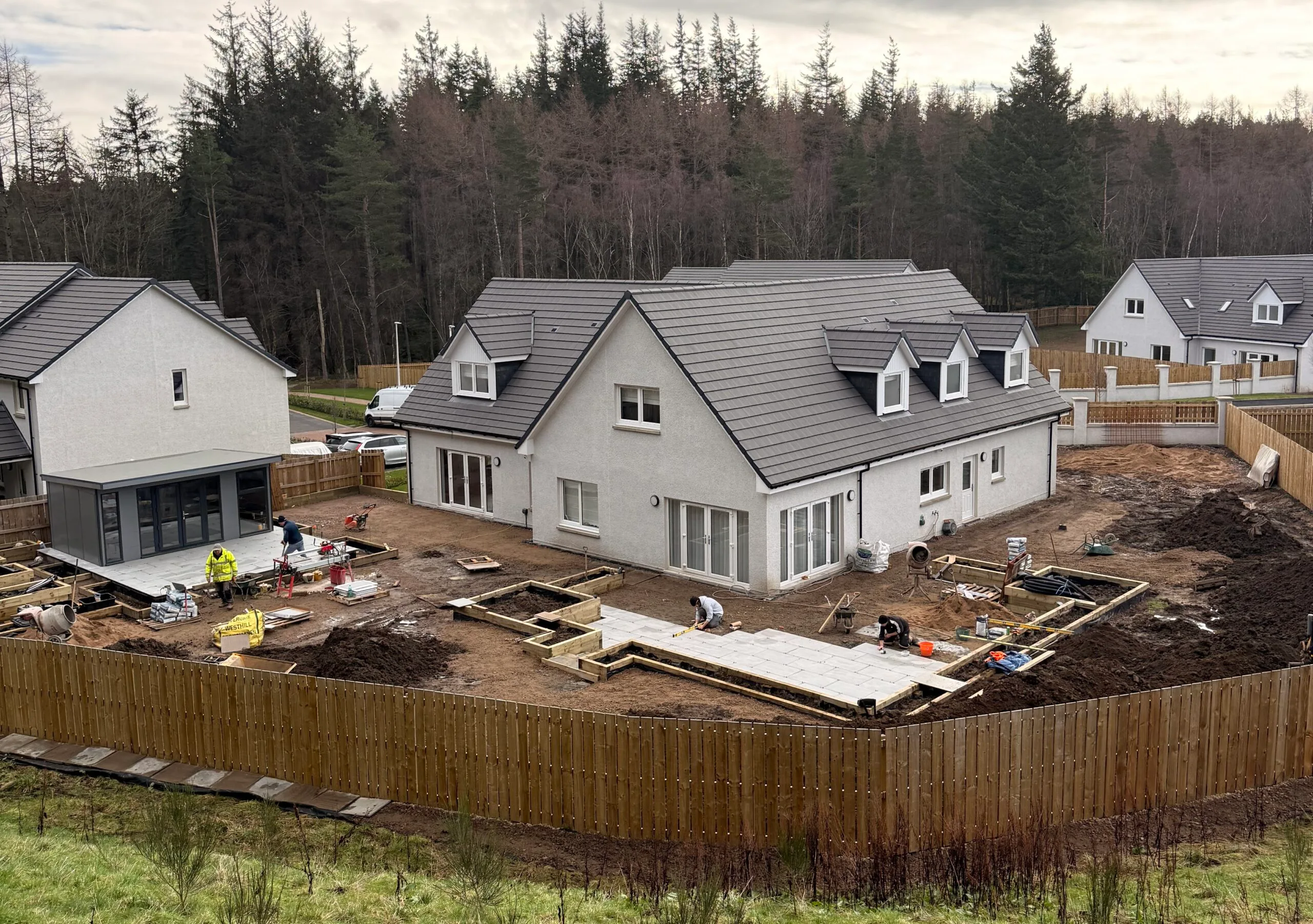 Construction site in a suburban neighborhood with new gray-roofed houses, workers laying foundations, and wooden fences around messy yards with a forest backdrop behind this row of homes.