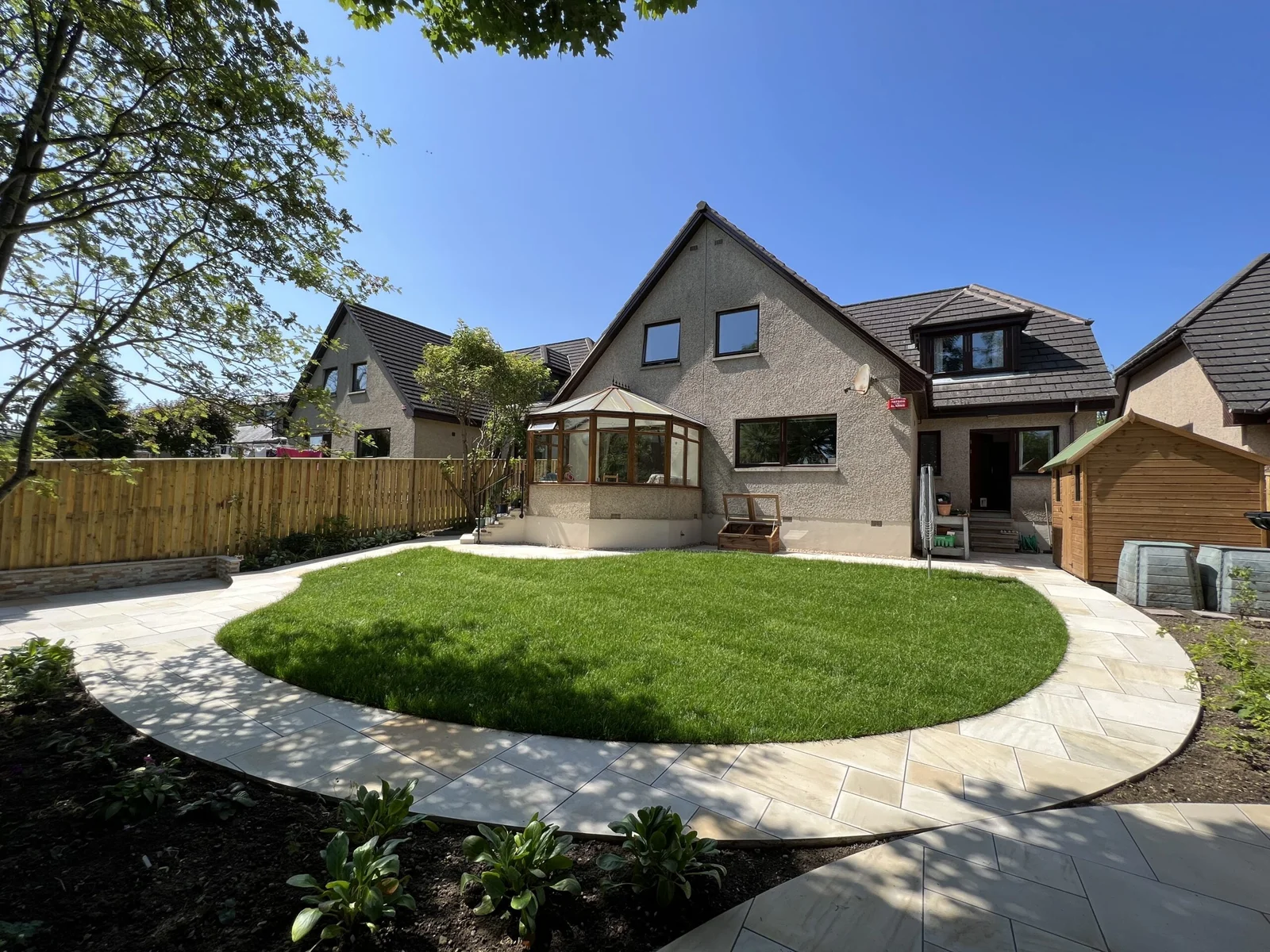 Backyard with a circular green lawn, light stone patio, wooden fence, and a beige house featuring a glass conservatory and small shed.