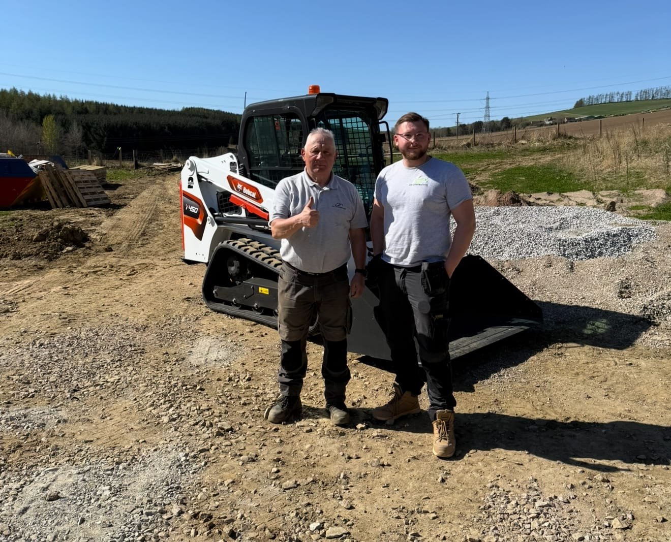 Two men stand beside a white and orange Bobcat skid-steer loader on a gravel construction site under a clear blue sky.