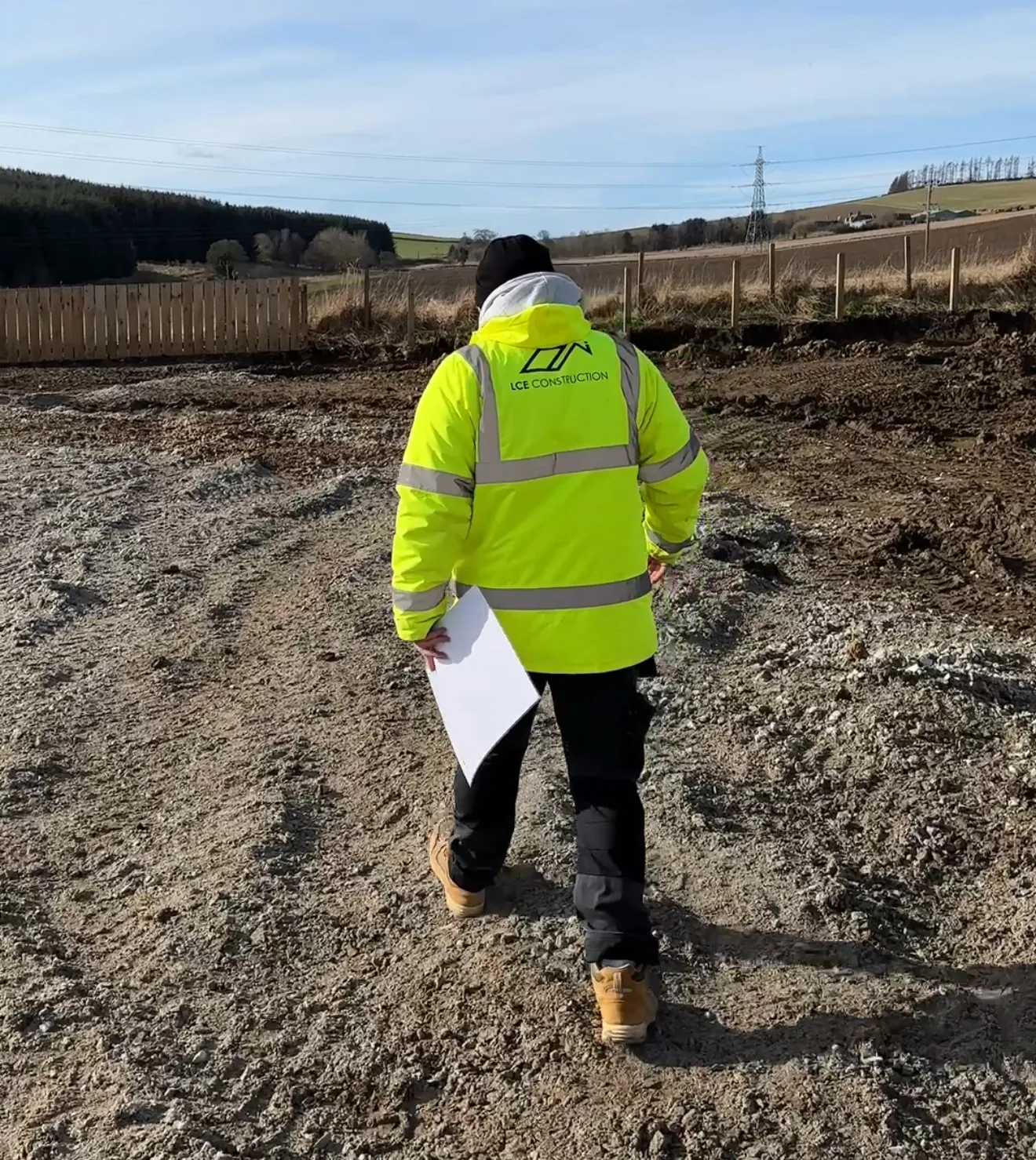 Back view of a construction worker in a neon high-visibility jacket with reflective stripes, holding a clipboard on a gravel site.