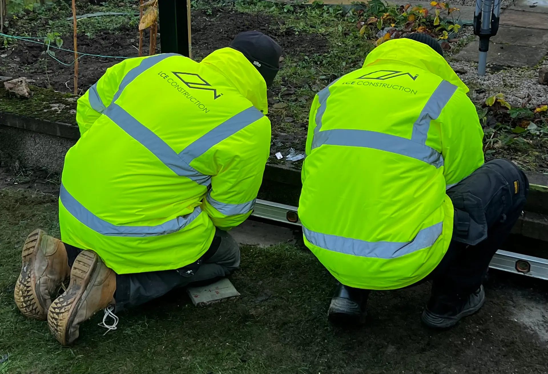 Two construction workers in neon yellow safety jackets crouch beside an open metal container outdoors, logo reading ICE CONSTRUCTION on their backs.