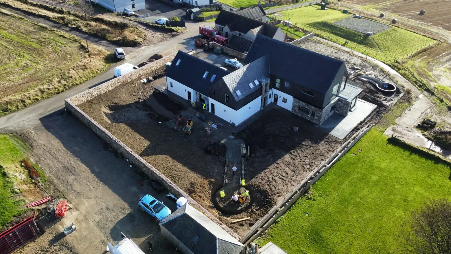 A white and dark-roofed house under construction with a large dirt yard, stone boundary wall, and workers around a circular foundation pit.