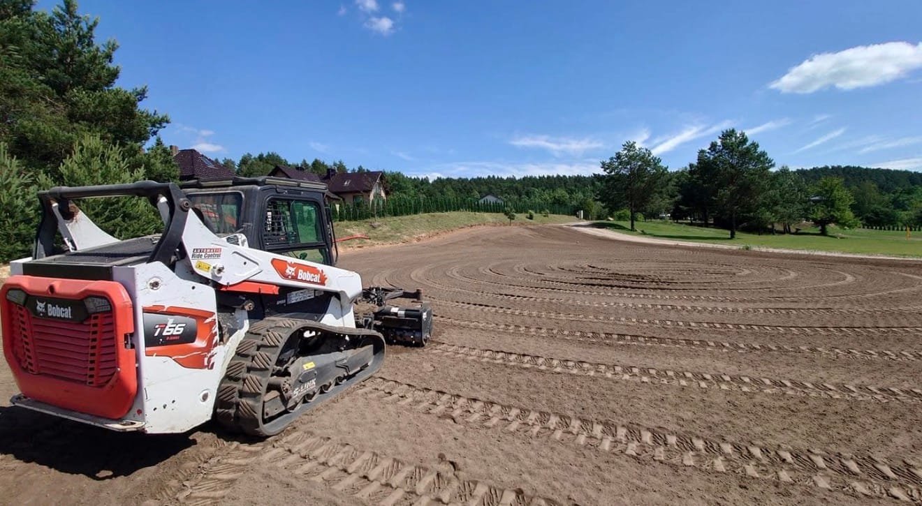 Compact tracked Bobcat loader on a freshly graded dirt lot, circular ruts visible in the soil with a blue sky above.