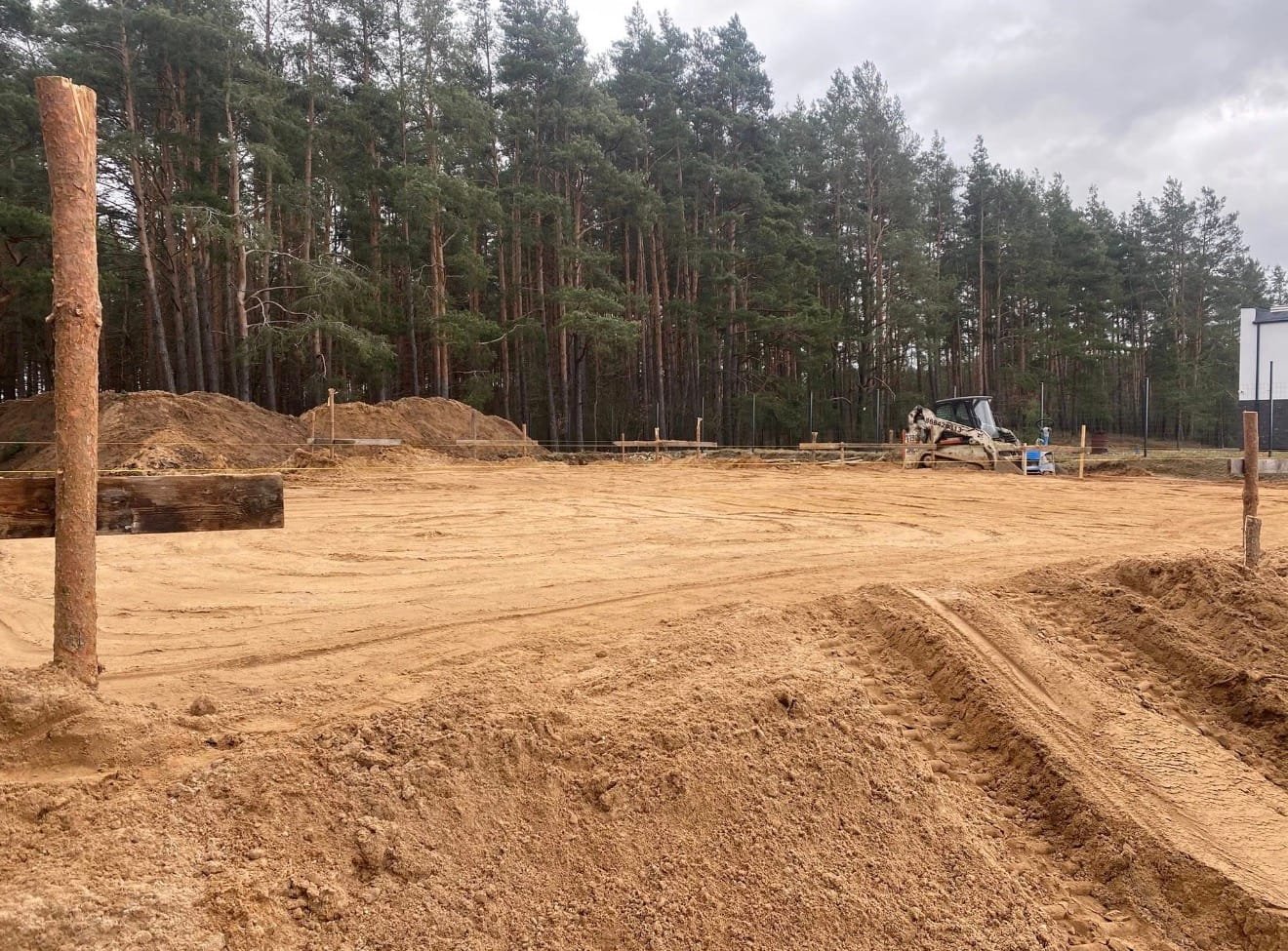 Cleared construction site with piles of dirt, wooden posts, and a small excavator against a backdrop of pine trees.