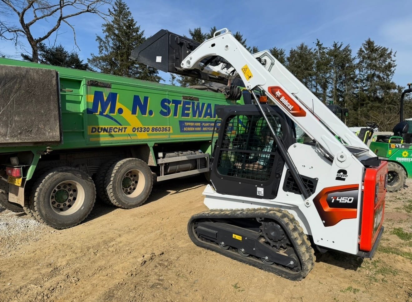 Compact Bobcat T450 excavator with tracked undercarriage beside a green dump truck on a dirt site, arm raised ready to load material.