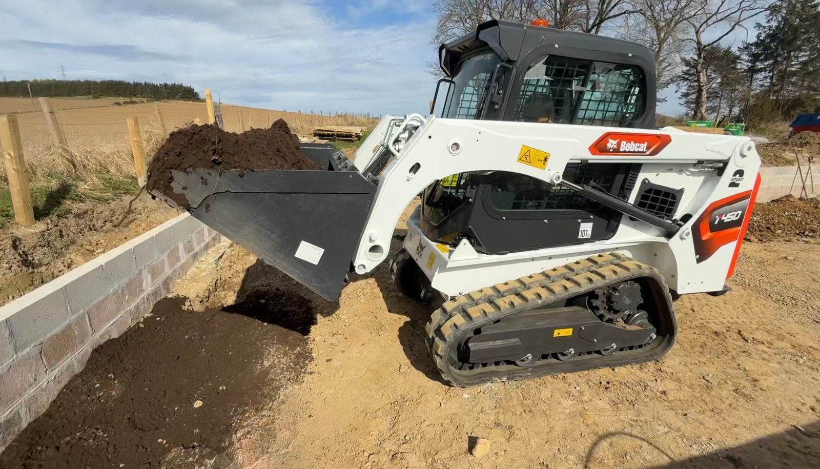 Bobcat skid-steer loader dumping dirt into a trench beside a low concrete wall on a construction site.