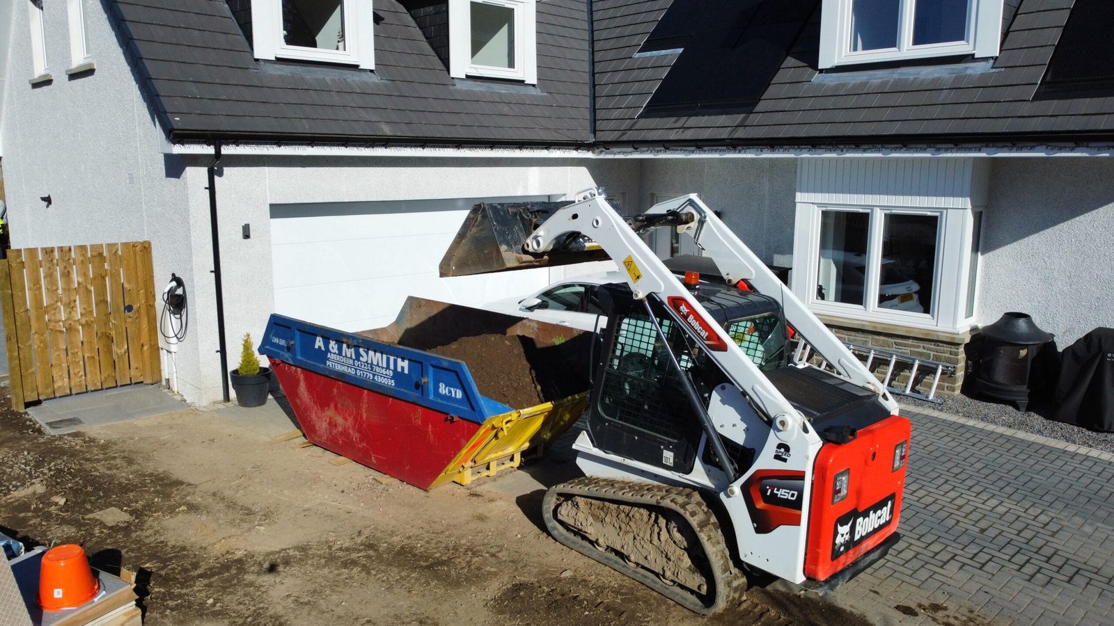 Residential construction scene with a Bobcat mini excavator loading dirt into a large skip in the driveway.