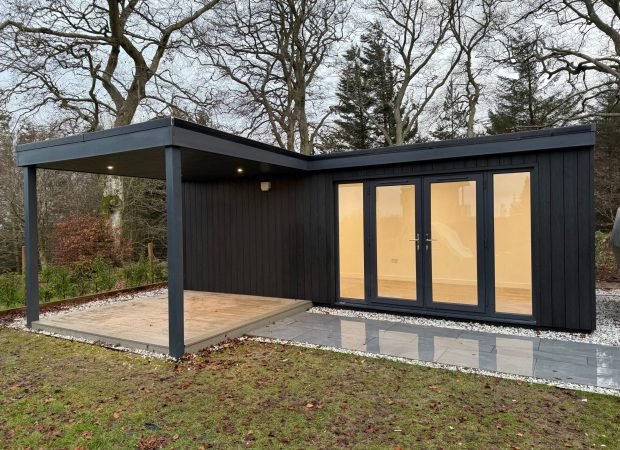 Dark blue modern garden shed with large glass doors and warm interior light, set against bare trees outdoors on a cloudy day.