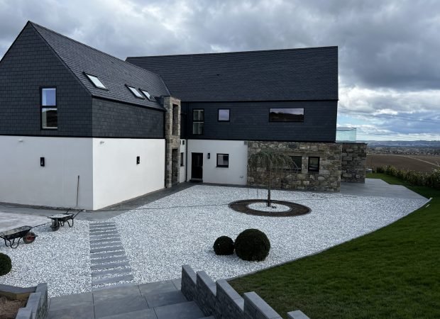 Modern dark-roofed house with white lower wall and stone accents, set in a gravel courtyard with stepping stones.